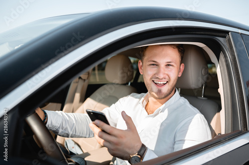 Young businessman smiling looking at mobile phone while driving a car. Man smiles lokking at the camera.
