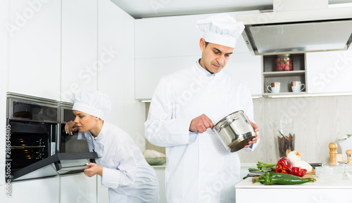 Professional chef in uniform with pot working with woman cook on kitchen