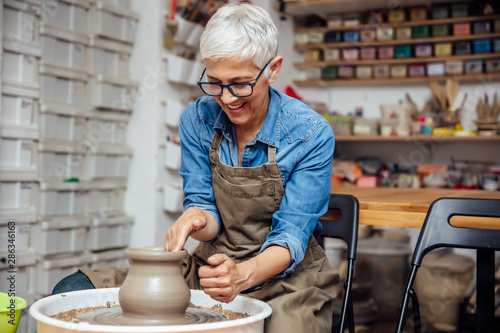 Senior female potter working on pottery wheel while sitting  in her workshop