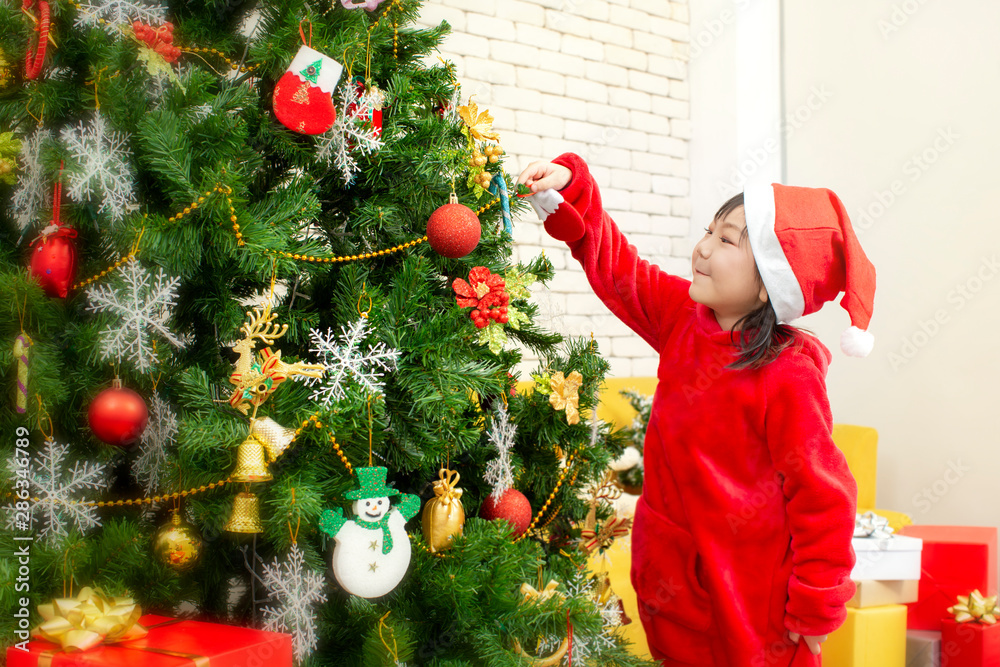 X-mas and holiday. The girl is hanging the decorative socks on the Christmas tree in the living room. Children in santa hat hand holding decoration. Cute little girl is decorating the christmas tree.