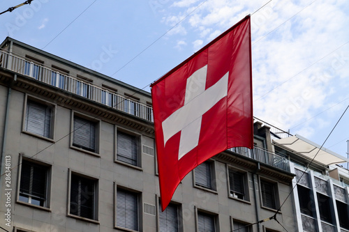 Flag of Switzerland hanging on middle of the street with building and blue cloudy sky in background 