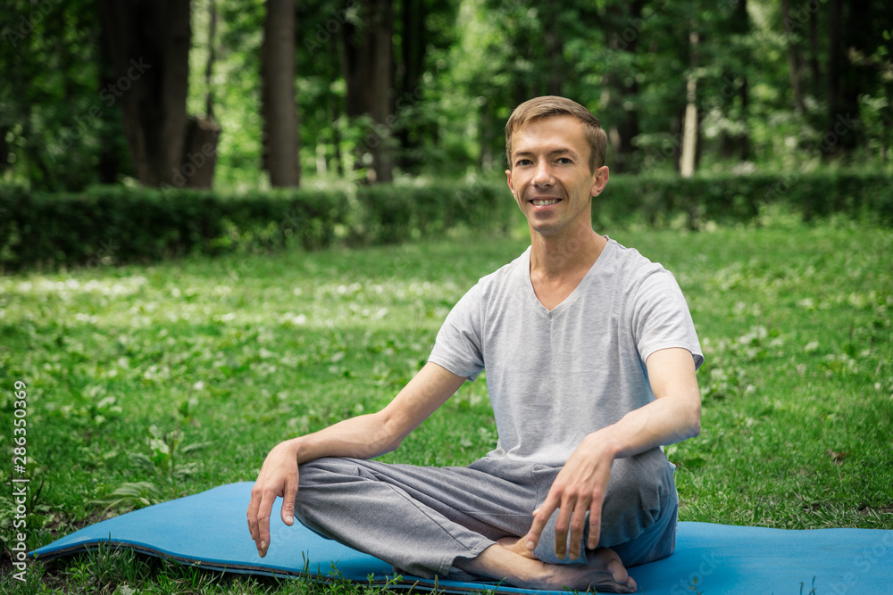 Obraz premium Young attractive man in sport clothes is meditating in the lotus position with a pacified face in the park against the background of green grass and trees.