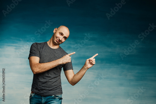 A young bald man in a gray T-shirt on a blue background fooling around and points to the side with two hands
