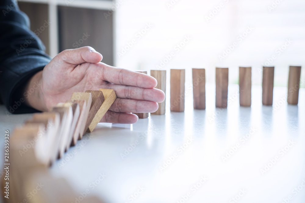 © Ngampol - Risk and Strategy in Business, Image of hand stopping falling collapse wooden block dominoes effect from continuous toppled block, prevention and development to stability
