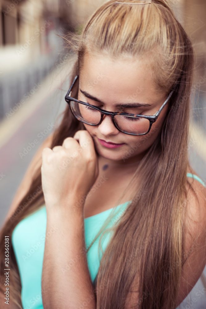Portrait of a young blonde girl with long hair and glasses, outdoors in the city. She is looking down like thoughtful Portrait of a young blonde girl with long hair and glasses, outdoors in the city. She is looking down like thoughtful