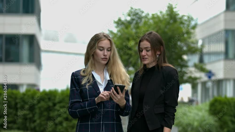 Two smartly dressed women look at a mobile device while chatting in a modern urban setting Two smartly dressed women look at a mobile device while chatting in a modern urban setting