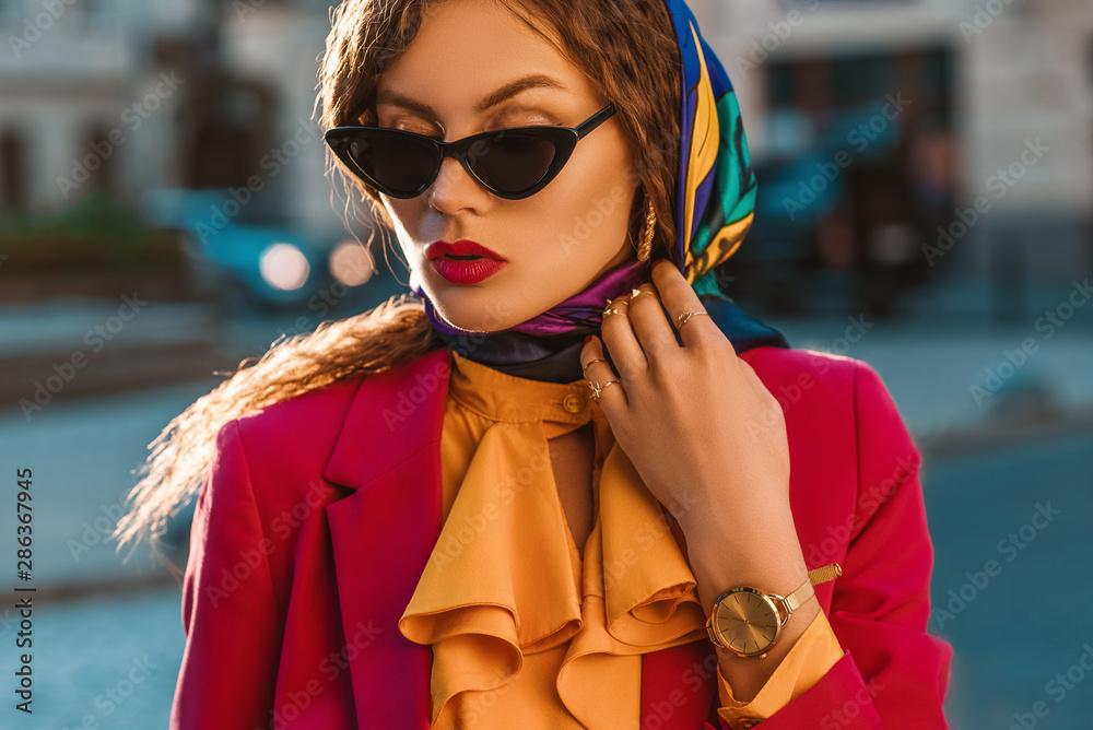 Outdoor close up fashion portrait elegant woman wearing colorful head scarf, black cat eye sunglasses, golden wrist watch, yellow blouse with jabot, pink blazer, walking in street of city Stock-foto