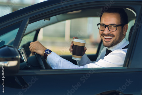 Smiling businessman driving a car while drinking coffee. He is looking at camera