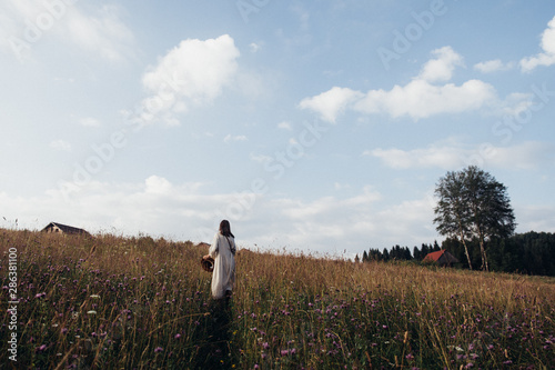 Trendy girl in stylish autumn dress with beautiful hat walking in the field with flowers and straw.