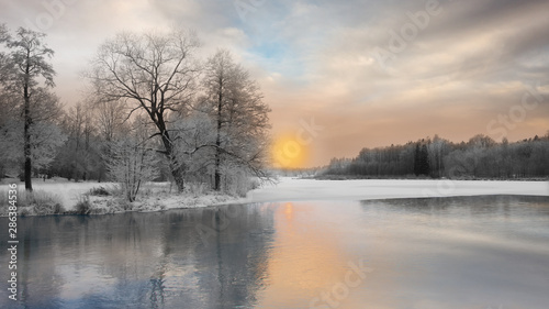Beautiful winter landscape with trees covered in rime frost, and reflection in water of sunset over river partly covered with ice © Pink Badger