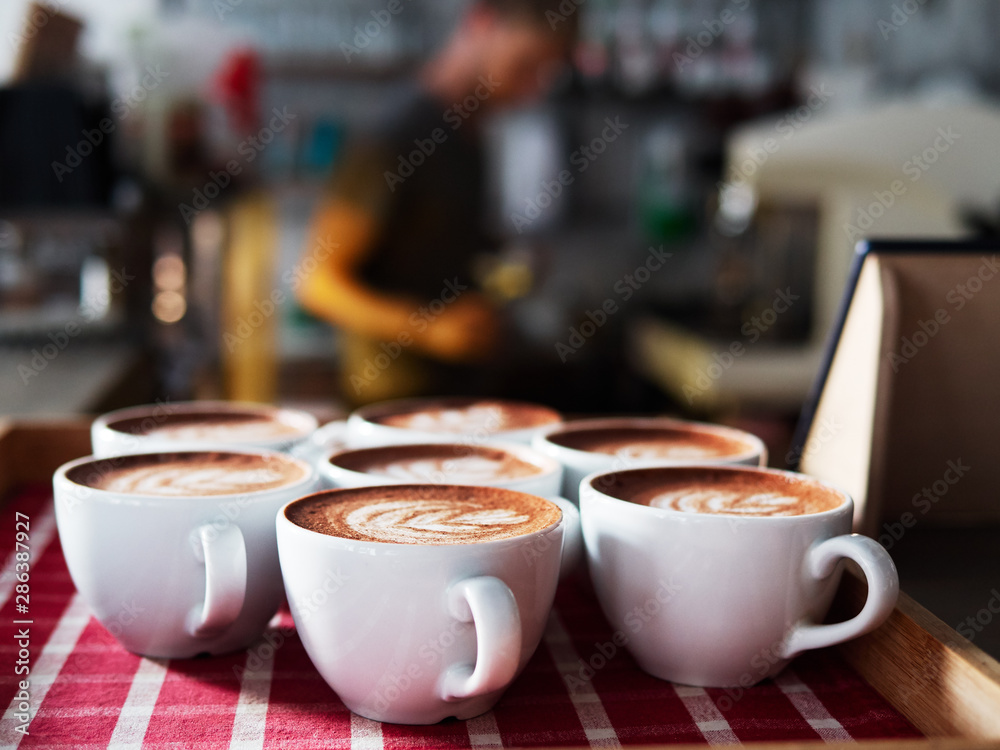 Set of freshly brewed cappuccino cups with milk foam latte art served on white red checkered tray in cozy city cafe lit with natural window light Set of freshly brewed cappuccino cups with milk foam latte art served on white red checkered tray in cozy city cafe lit with natural window light