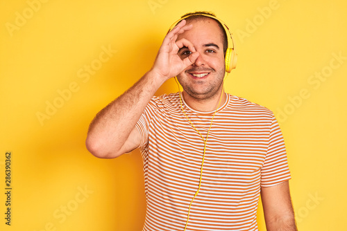 Young man listening to music using headphones standing over isolated yellow background doing ok gesture with hand smiling, eye looking through fingers with happy face. © Krakenimages.com