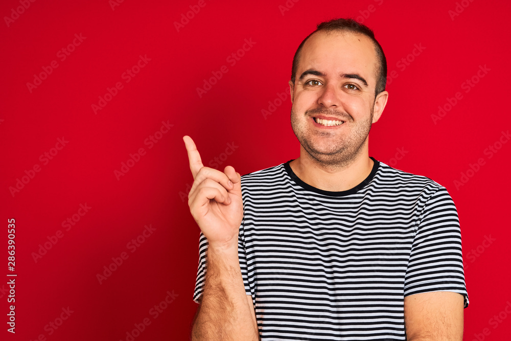 Naklejka premium Young man wearing striped navy t-shirt standing over isolated red background with a big smile on face, pointing with hand and finger to the side looking at the camera.