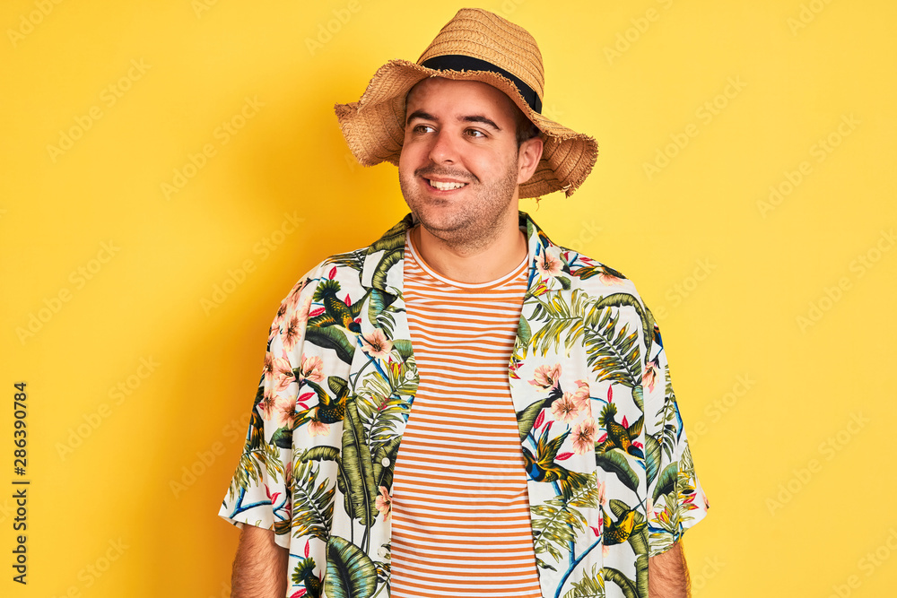 Young man on vacation wearing summer shirt and hat over isolated yellow background looking away to side with smile on face, natural expression. Laughing confident. Young man on vacation wearing summer shirt and hat over isolated yellow background looking away to side with smile on face, natural expression. Laughing confident.