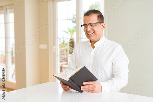 Middle age man reading a book at home wearing glasses with a happy face standing and smiling with a confident smile showing teeth