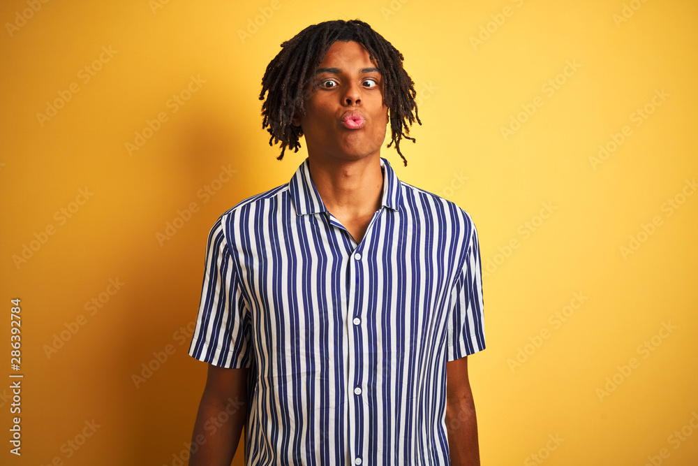Afro man with dreadlocks wearing casual striped t-shirt over isolated yellow background making fish face with lips, crazy and comical gesture. Funny expression.