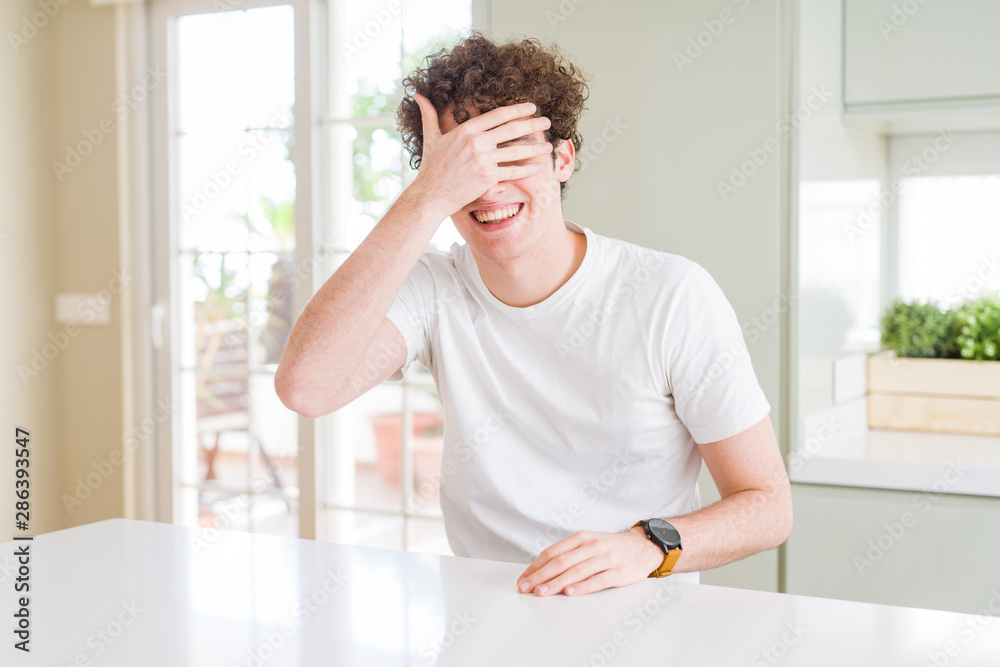 Young handsome man wearing white t-shirt at home smiling and laughing with hand on face covering eyes for surprise. Blind concept. Young handsome man wearing white t-shirt at home smiling and laughing with hand on face covering eyes for surprise. Blind concept.