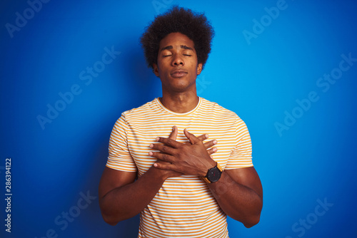 American man with afro hair wearing striped yellow t-shirt over isolated blue background smiling with hands on chest with closed eyes and grateful gesture on face. Health concept. © Krakenimages.com