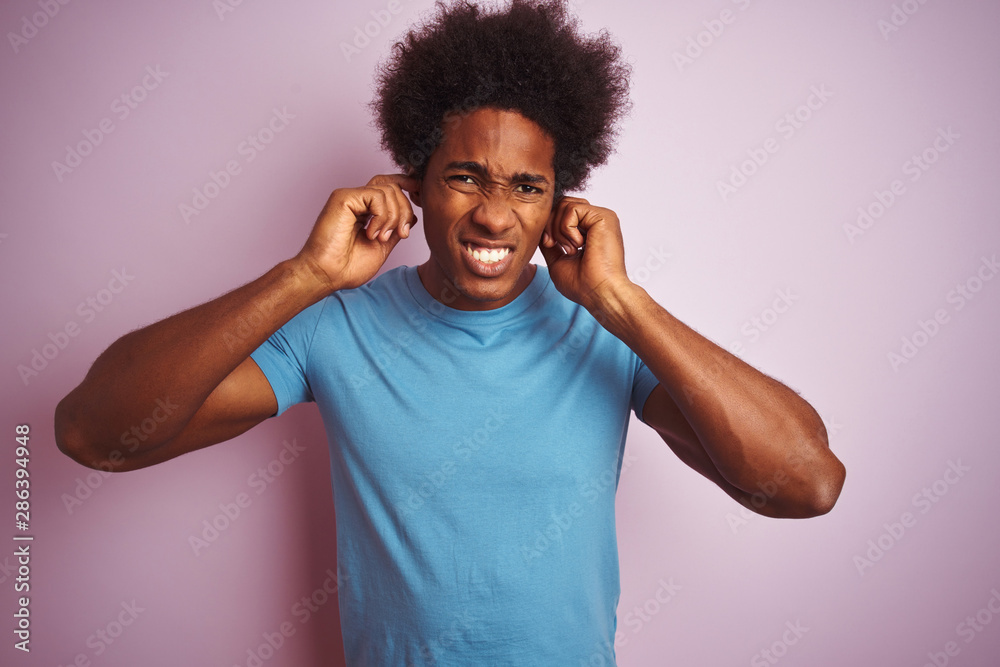 African american man with afro hair wearing blue t-shirt standing over isolated pink background covering ears with fingers with annoyed expression for the noise of loud music. Deaf concept. African american man with afro hair wearing blue t-shirt standing over isolated pink background covering ears with fingers with annoyed expression for the noise of loud music. Deaf concept.