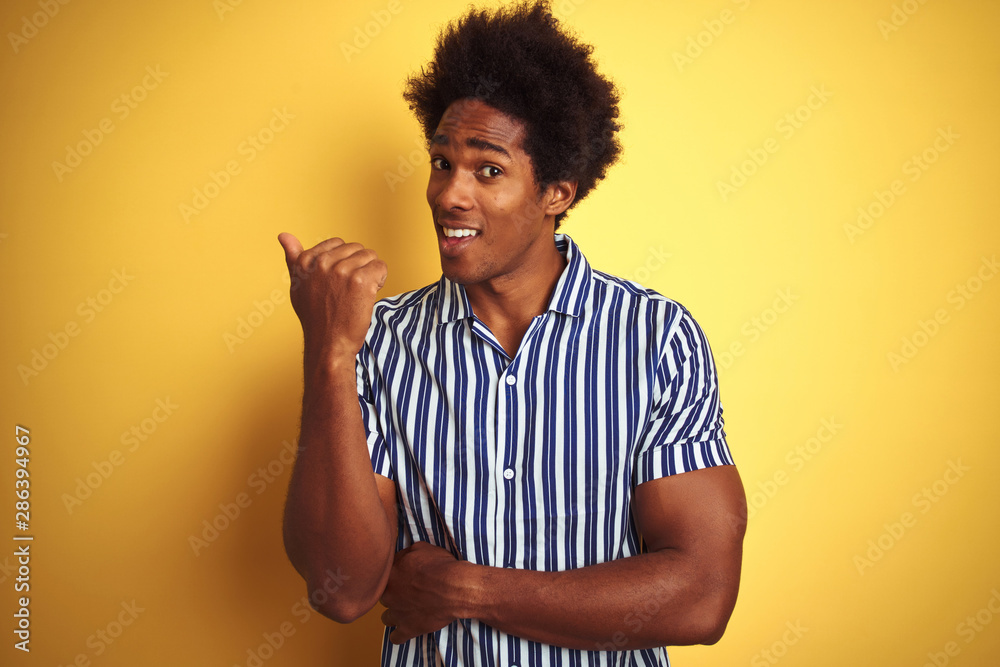 American man with afro hair wearing striped shirt standing over isolated yellow background smiling with happy face looking and pointing to the side with thumb up. American man with afro hair wearing striped shirt standing over isolated yellow background smiling with happy face looking and pointing to the side with thumb up.