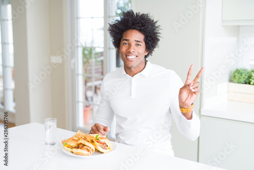 African American hungry man eating hamburger for lunch smiling with happy face winking at the camera doing victory sign. Number two.