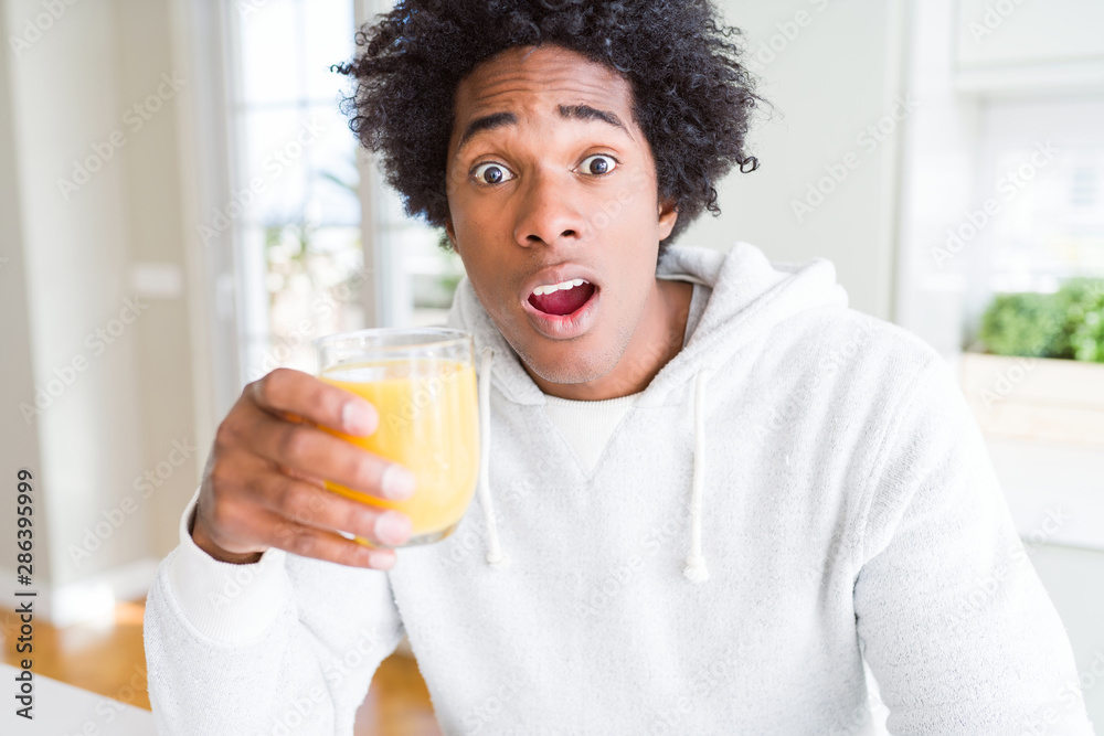 African American man holding and drinking glass of orange juice scared in shock with a surprise face, afraid and excited with fear expression African American man holding and drinking glass of orange juice scared in shock with a surprise face, afraid and excited with fear expression