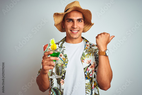 Young handsome man drinking cocktail on holiday over white isolated background pointing and showing with thumb up to the side with happy face smiling