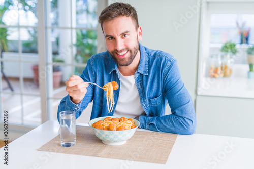 Handsome man eating pasta with meatballs and tomato sauce at home while smiling at the camera