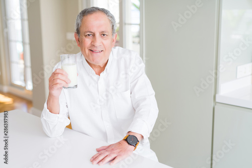 Handsome senior man drinking a glass of fresh milk at breakfast with a happy face standing and smiling with a confident smile showing teeth