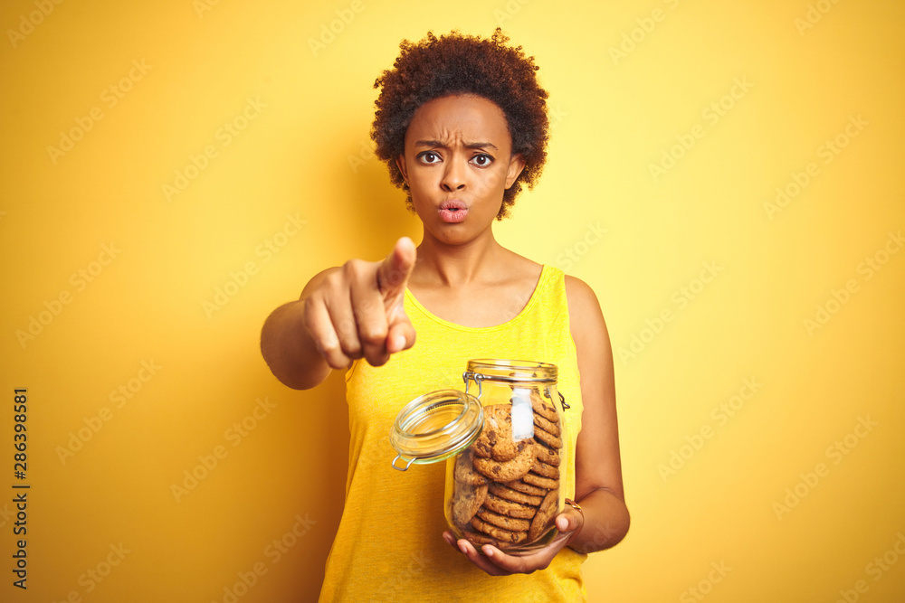 African american woman holding jar of chocolate chips cookies over yellow background pointing with finger to the camera and to you, hand sign, positive and confident gesture from the front African american woman holding jar of chocolate chips cookies over yellow background pointing with finger to the camera and to you, hand sign, positive and confident gesture from the front