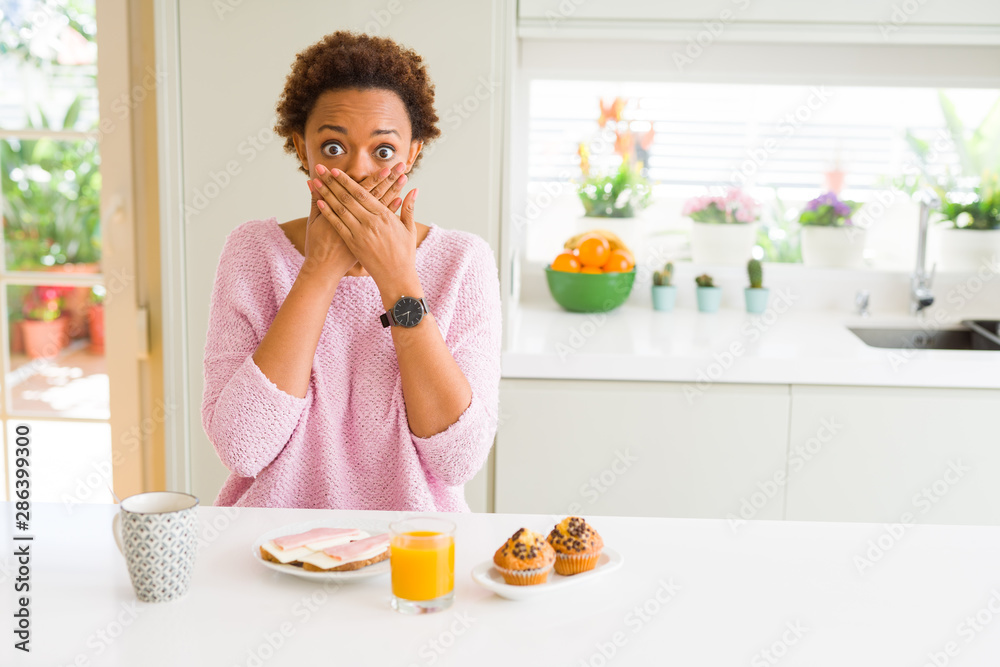 Young african american woman eating breaksfast in the morning at home shocked covering mouth with hands for mistake. Secret concept.