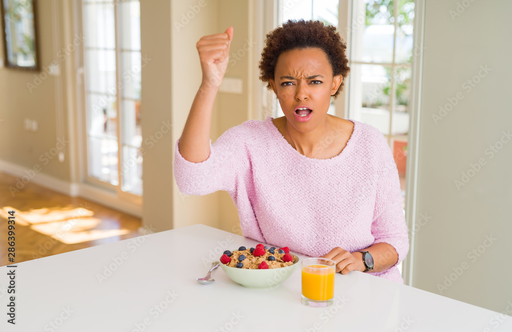 Young african american woman having healthy breakfast in the morning at home angry and mad raising fist frustrated and furious while shouting with anger. Rage and aggressive concept. Young african american woman having healthy breakfast in the morning at home angry and mad raising fist frustrated and furious while shouting with anger. Rage and aggressive concept.