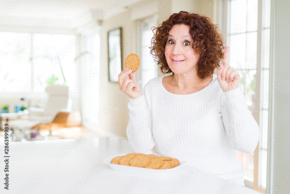 Senior woman eating healthy whole grain biscuit at home surprised with an idea or question pointing finger with happy face, number one Senior woman eating healthy whole grain biscuit at home surprised with an idea or question pointing finger with happy face, number one