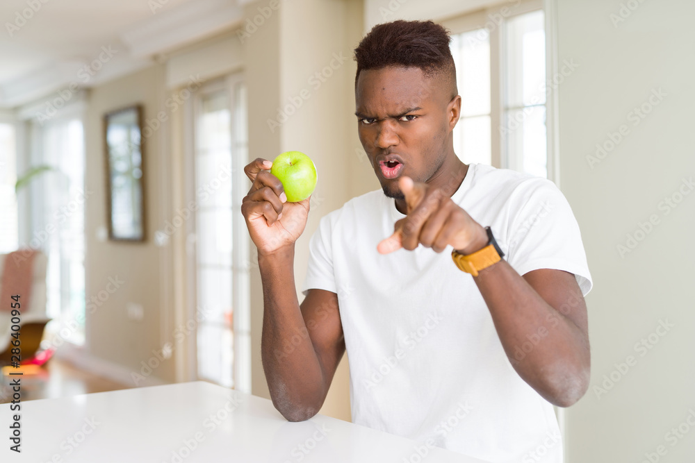 Young african american man eating fresh green apple pointing with finger to the camera and to you, hand sign, positive and confident gesture from the front Young african american man eating fresh green apple pointing with finger to the camera and to you, hand sign, positive and confident gesture from the front