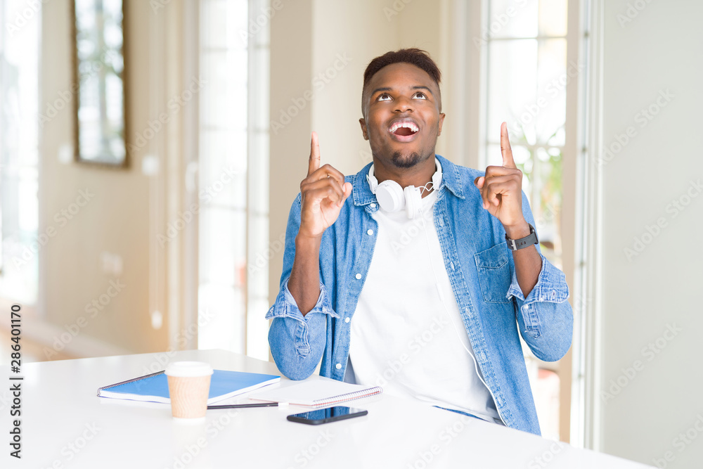 African american student man studying using notebooks and wearing headphones amazed and surprised looking up and pointing with fingers and raised arms. African american student man studying using notebooks and wearing headphones amazed and surprised looking up and pointing with fingers and raised arms.