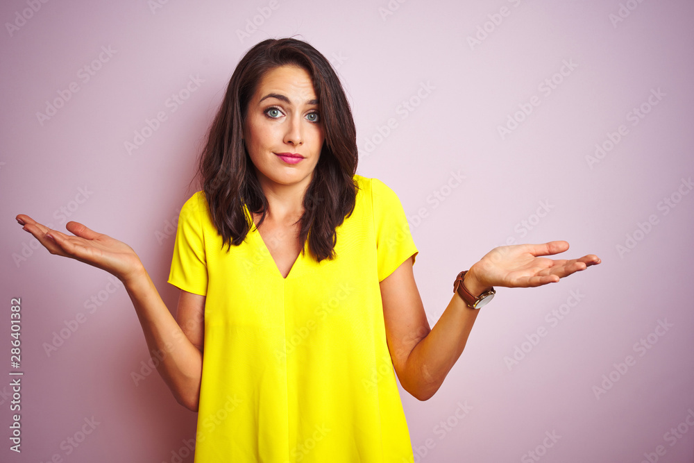 Young beautiful woman wearing yellow t-shirt standing over pink isolated background clueless and confused expression with arms and hands raised. Doubt concept. Young beautiful woman wearing yellow t-shirt standing over pink isolated background clueless and confused expression with arms and hands raised. Doubt concept.