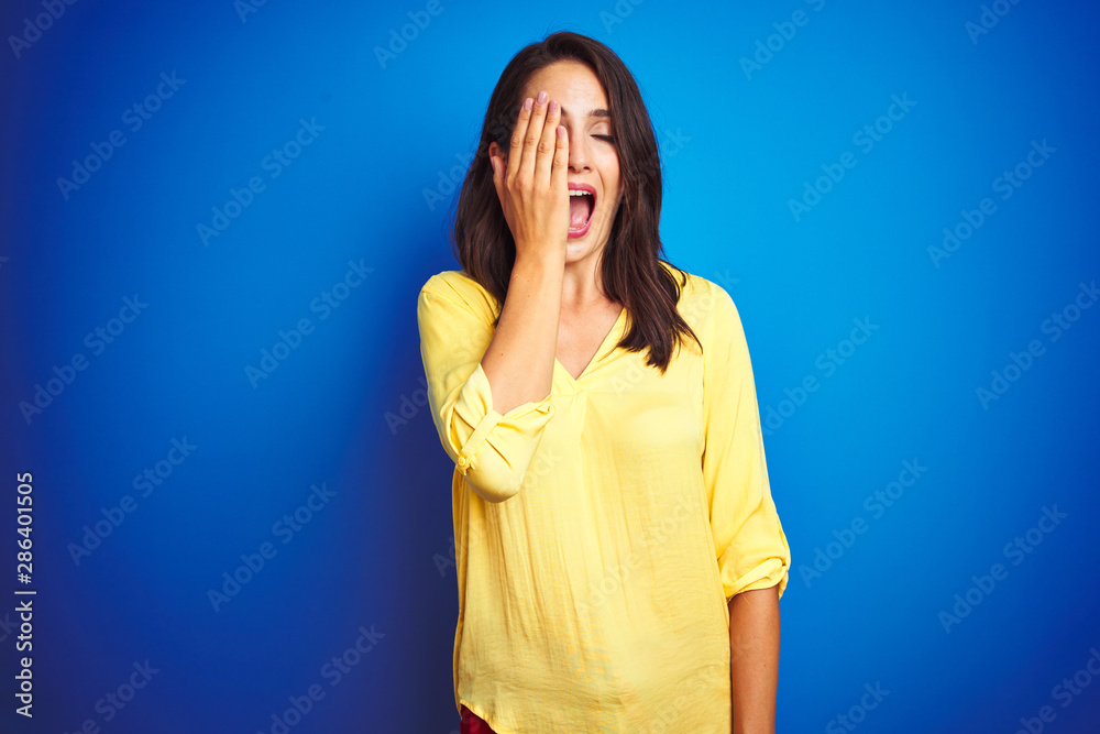 Young beautiful woman wearing yellow t-shirt standing over blue isolated background covering one eye with hand, confident smile on face and surprise emotion. Young beautiful woman wearing yellow t-shirt standing over blue isolated background covering one eye with hand, confident smile on face and surprise emotion.