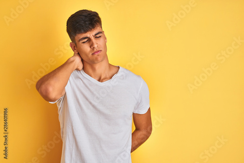 Young indian man wearing white t-shirt standing over isolated yellow background Suffering of neck ache injury, touching neck with hand, muscular pain