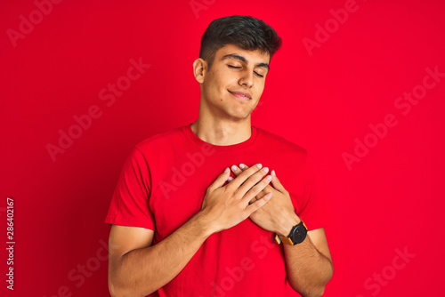 Young indian man wearing t-shirt standing over isolated red background smiling with hands on chest with closed eyes and grateful gesture on face. Health concept.