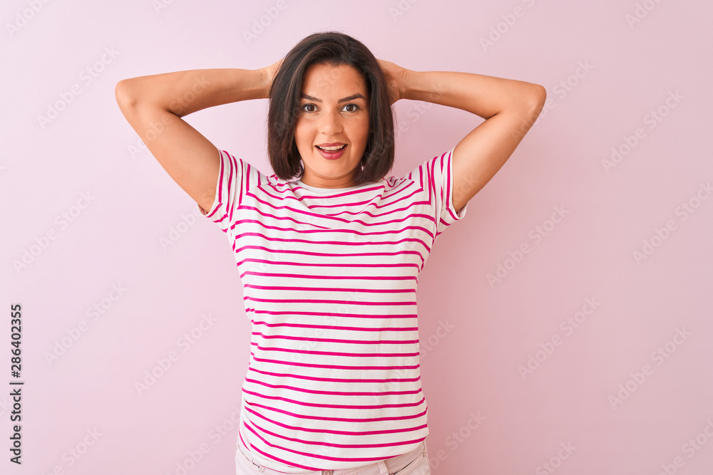 Young beautiful woman wearing striped t-shirt standing over isolated pink background relaxing and stretching, arms and hands behind head and neck smiling happy