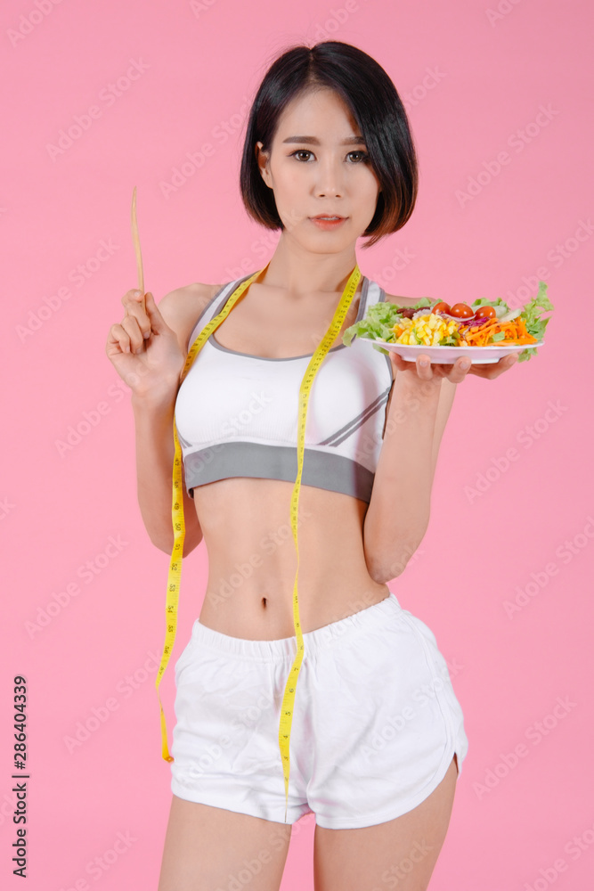 Healthy woman standing holdding vegetable salad diet food and tape measure in home on pink backgrounds, Healthy concept, Diet concept Healthy woman standing holdding vegetable salad diet food and tape measure in home on pink backgrounds, Healthy concept, Diet concept