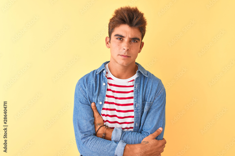 Young handsome man wearing striped t-shirt and denim shirt over isolated yellow background skeptic and nervous, disapproving expression on face with crossed arms. Negative person. Young handsome man wearing striped t-shirt and denim shirt over isolated yellow background skeptic and nervous, disapproving expression on face with crossed arms. Negative person.