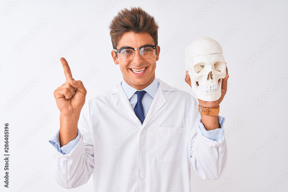 Young handsome doctor man wearing glasses holding skull over isolated white background very happy pointing with hand and finger to the side Young handsome doctor man wearing glasses holding skull over isolated white background very happy pointing with hand and finger to the side