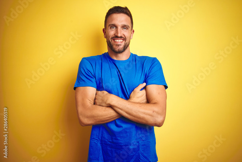 Young handsome man wearing casual blue t-shirt over yellow isolated background happy face smiling with crossed arms looking at the camera. Positive person.