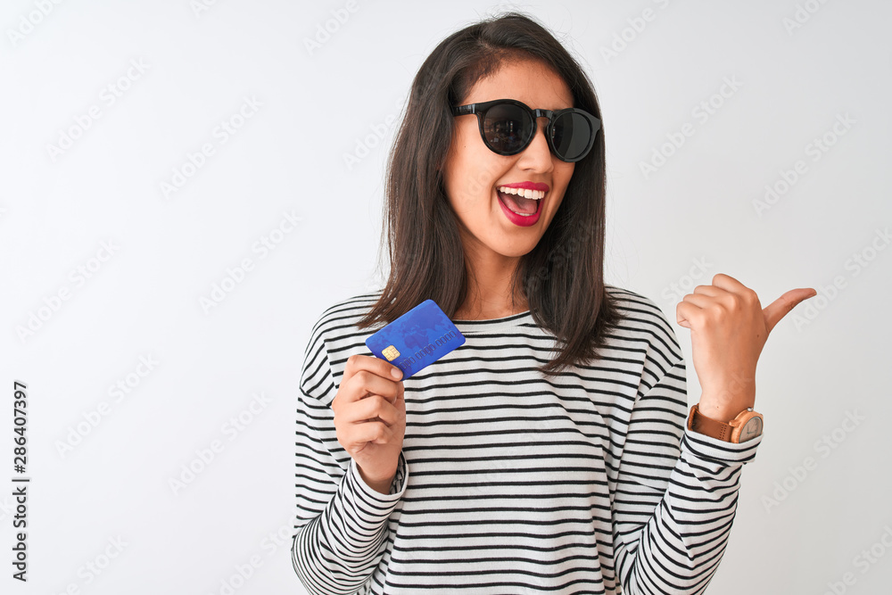 Young chinese woman wearing sunglasses holding credit card over isolated white background pointing and showing with thumb up to the side with happy face smiling Young chinese woman wearing sunglasses holding credit card over isolated white background pointing and showing with thumb up to the side with happy face smiling