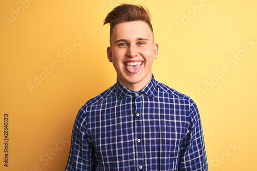 Young handsome man wearing casual shirt standing over isolated yellow background sticking tongue out happy with funny expression. Emotion concept.