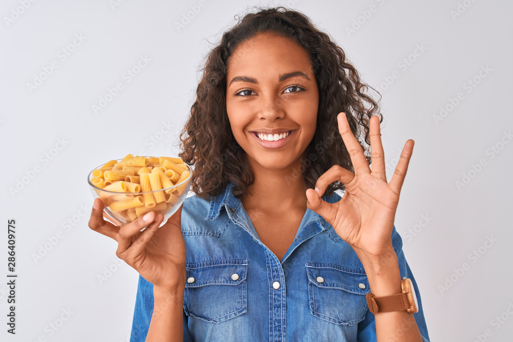 Young brazilian woman holding bowl with macaroni pasta over isolated white background doing ok sign with fingers, excellent symbol Young brazilian woman holding bowl with macaroni pasta over isolated white background doing ok sign with fingers, excellent symbol