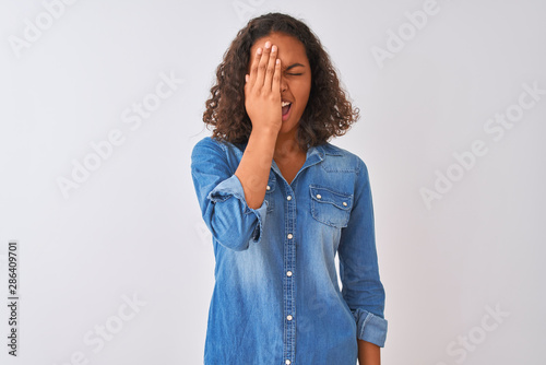 Young brazilian woman wearing denim shirt standing over isolated white background Yawning tired covering half face, eye and mouth with hand. Face hurts in pain.