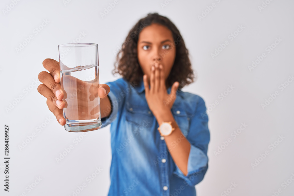 Young brazilian woman holding glass of water standing over isolated white background cover mouth with hand shocked with shame for mistake, expression of fear, scared in silence, secret concept Young brazilian woman holding glass of water standing over isolated white background cover mouth with hand shocked with shame for mistake, expression of fear, scared in silence, secret concept