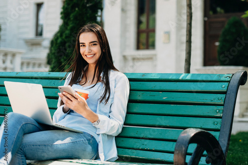 Portrait of a charming long haired girl sitting on a beach and while using a smartphone looking at camera laughing against a building with a laptop on her legs.